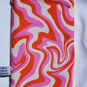 May include: A rectangular zipped pouch featuring a retro wavy design in pink, orange, and off-white. The pouch has a matching orange zip and a small white tag with the text "while sadie sleeps".
