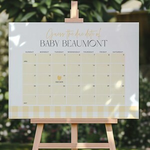 May include: A baby shower sign on a wooden easel. The sign reads "Guess the due date of Baby Beaumont" above a calendar grid. The calendar is light yellow with a gingham border. The background is a blurred outdoor scene.