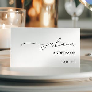 May include: A white place card with the name "Juliana" in elegant script, "Andersson" in a serif font, and "Table 1" below. The card sits on a white plate, with a candle and glassware in the background.