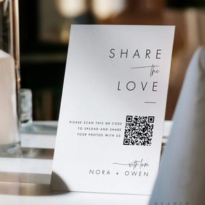 May include: A white wedding sign with the text "SHARE THE LOVE" and a QR code, next to lit candles in glass holders. The sign also includes the names "NORA + OWEN".