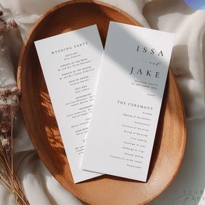 以下が含まれることがあります： IssaとJakeの名前、2045年9月20日の日付、結婚式のプログラムが記載された、黒いセリフ体の文字が印刷された2枚の白いウェディングプログラムカード。カードは滑らかな楕円形の木製トレイの上に置かれ、青いCanva編集バッジが付いています。