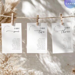 May include: Three white wedding table cards are suspended from a string with wooden clothespins. Each card is labeled "Table One," "Table Two," and "Table Three" in elegant script, with guest names listed below. The design is minimalist and modern.