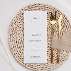 May include: A white card with the text "welcome let's eat!" on a woven placemat with gold silverware and a pink napkin. The menu includes Mediterranean Tossed Salad, Honey Herb Roasted Chicken, Grilled Pork Tenderloin, and Specialty Wedding Cake.