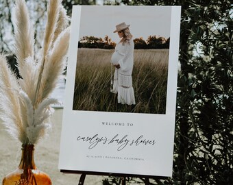May include: Welcome to Carly's Baby Shower sign with a photo of a pregnant woman in a white dress and hat standing in a field of tall grass. The sign is on a wooden easel.