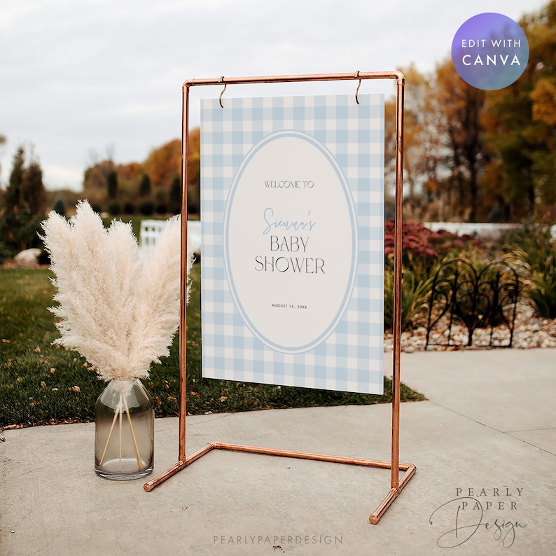 May include: A copper-colored metal frame holds a baby shower sign with a light blue gingham pattern. The sign reads "Welcome to Sunny's Baby Shower" in elegant script. A vase of fluffy, dried pampas grass sits to the left of the sign.