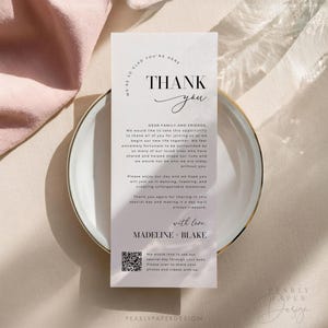 Op de afbeelding: Een witte bedankkaart met een zwarte rand en de tekst "We're so glad you're here. Thank you." De kaart bevat ook een bericht waarin de gasten worden bedankt voor hun aanwezigheid op de bruiloft en een QR-code om foto's en video's te delen. De kaart is op een marmeren achtergrond.