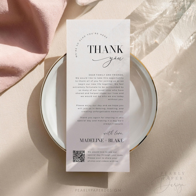 Op de afbeelding: Een witte bedankkaart met een zwarte rand en de tekst "We're so glad you're here. Thank you." De kaart bevat ook een bericht waarin de gasten worden bedankt voor hun aanwezigheid op de bruiloft en een QR-code om foto's en video's te delen. De kaart is op een marmeren achtergrond.