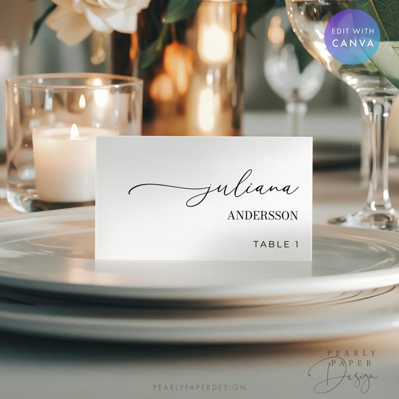May include: A white place card with the name "Juliana" in elegant script, "Andersson" in a serif font, and "Table 1" below. The card sits on a white plate, with a candle and glassware in the background.