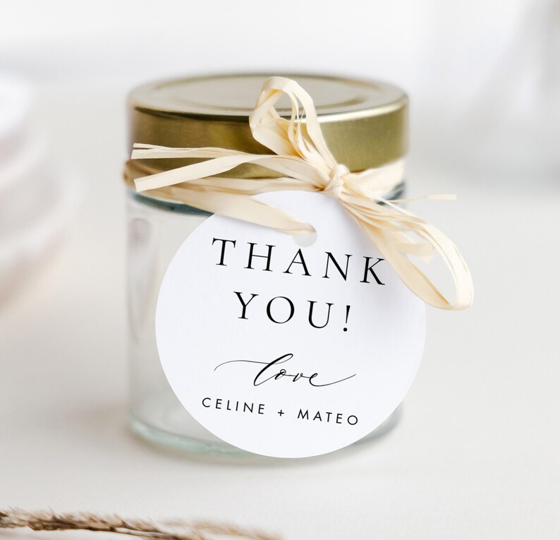 May include: White round thank you tag with the text "Thank You! Love Celine + Mateo" tied to a clear glass jar with a gold lid and a beige ribbon.