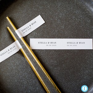 May include: Two gold straws with white labels that say "STELLA & RYAN 08.08.2020" on a dark gray surface.