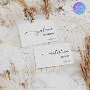 May include: Two white place cards with elegant black calligraphy. One card reads "juliana ANDERSSON TABLE 1", the other reads "sebastian JAMESON TABLE 1". The cards are set on a textured white surface.