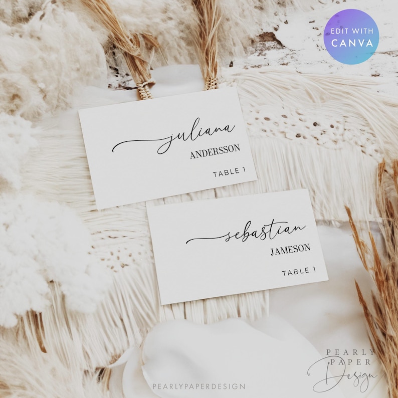 May include: Two white place cards with elegant black calligraphy. One card reads "juliana ANDERSSON TABLE 1", the other reads "sebastian JAMESON TABLE 1". The cards are set on a textured white surface.
