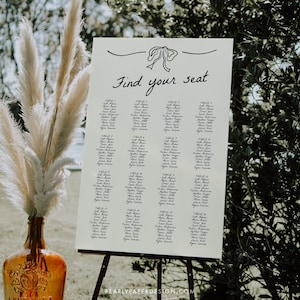 May include: A white seating chart with black calligraphy writing. The chart is titled "Find your seat" and lists guest names by table number. The chart is displayed on an easel.