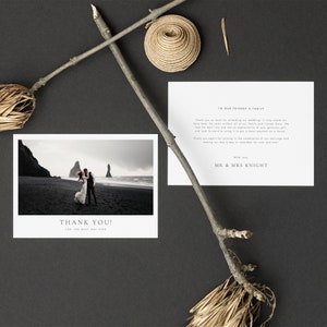 May include: A wedding invitation suite featuring a black and white photograph of a couple on a beach, a thank you card, and a card addressed to friends and family. The cards are accompanied by decorative twigs and a woven element, all set against a dark background.