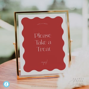 May include: A gold-framed sign with a red scalloped border and white text that reads "Love is Sweet. Please Take a Treat. Enjoy!" The sign is sitting on a wooden table.