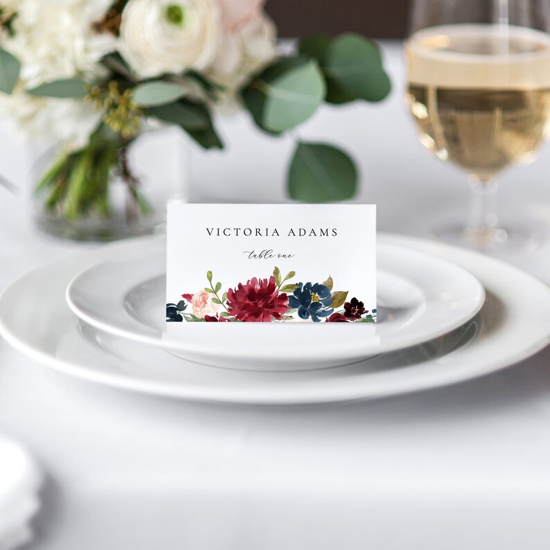 May include: A white place card with the text "VICTORIA ADAMS - table one" and a watercolour floral design featuring burgundy, blue, and pink flowers.