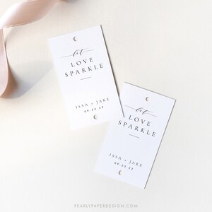 May include: Two white rectangular tags with the words "let LOVE SPARKLE" in elegant black script. Below, it reads "ISSA + JAKE 09.25.22". Each tag has a small hole at the top. A pale pink ribbon lies in the background.