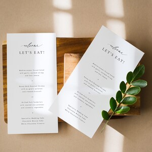 May include: Two white cards with the text "welcome let's eat!" on them. The cards are on a wooden surface with a green plant leaf.