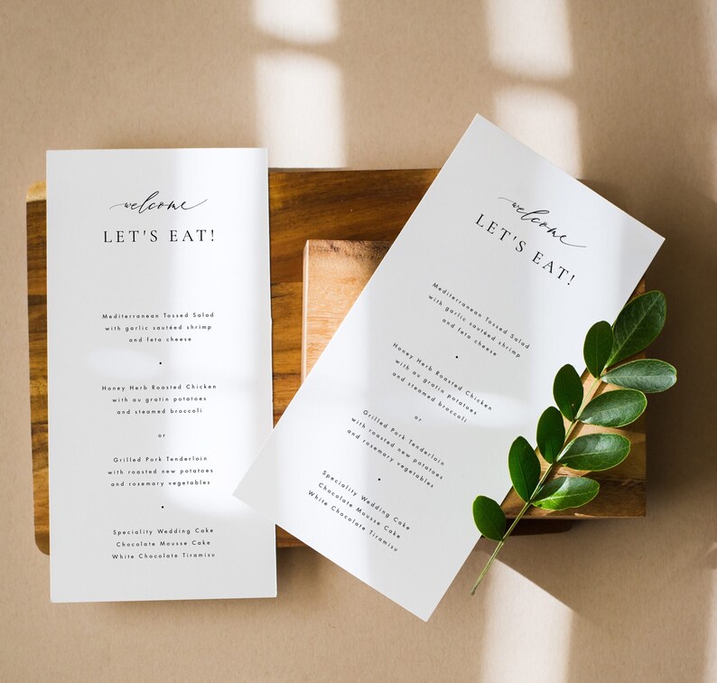 May include: Two white cards with the text "welcome let's eat!" on them. The cards are on a wooden surface with a green plant leaf.