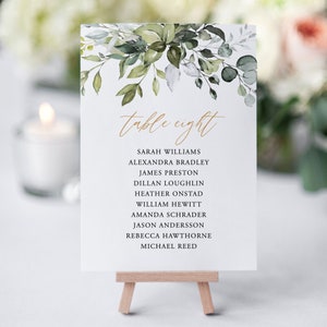 May include: A white table number card with a watercolour floral design in shades of green and grey. The card reads "table eight" in gold script and lists the names of guests assigned to this table.
