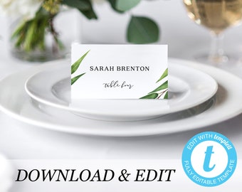 May include: A white place card with the name "SARAH BRENTON" and "table four" in elegant script. The card is decorated with green eucalyptus leaves and sits on a white plate. The words "DOWNLOAD & EDIT" are at the bottom.