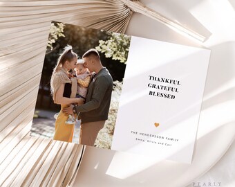 May include: A white card with black text that reads "Thankful Grateful Blessed" and a small orange heart. Below the text is "The Henderson Family, Emmy, Olivia and Carl". A photo of a family of three is in the background.