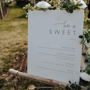 Puede incluir: Un cartel blanco con texto negro que dice "Love is Sweet" y enumera pastel de bodas, tiramis&uacute; de chocolate, cupcakes y donas. El cartel est&aacute; apoyado en un caballete de madera.