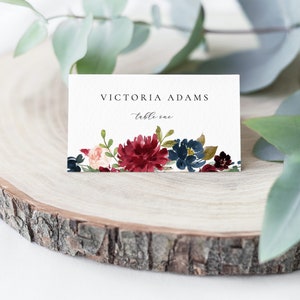 May include: White wedding place card with burgundy and blue watercolour floral design. The card reads "Victoria Adams, table one."