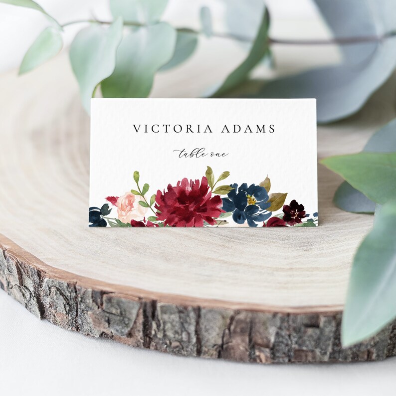 May include: White wedding place card with burgundy and blue watercolour floral design. The card reads "Victoria Adams, table one."