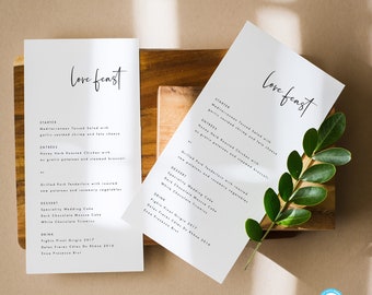 May include: Two white cards with the text "Love feast" printed in black calligraphy. The cards are laid out on a wooden surface and feature a wedding menu with starter, entree, dessert, and drink options.