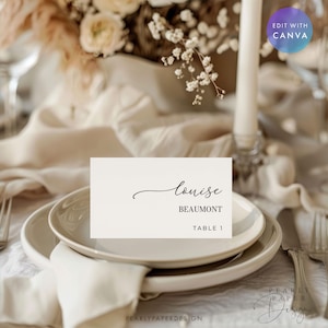 May include: A place setting with a white card featuring the name "Louise" in elegant script, with "Beaumont" below. The card is on a white plate, with a napkin and silverware. The background includes flowers and a candle.