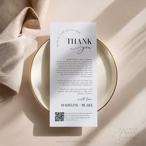 Op de afbeelding: Een witte bedankkaart met zwarte tekst. De kaart zegt "We're so glad you're here. Thank you." Het bevat ook een bericht aan "Dear Family and Friends" en een QR-code. De kaart ligt op een beige oppervlak.