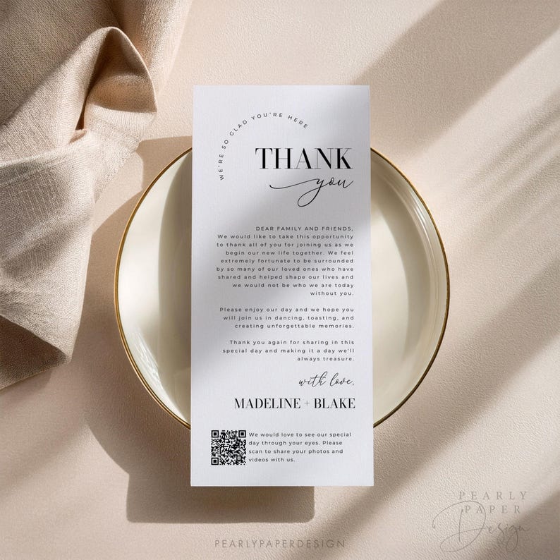 Op de afbeelding: Een witte bedankkaart met zwarte tekst. De kaart zegt "We're so glad you're here. Thank you." Het bevat ook een bericht aan "Dear Family and Friends" en een QR-code. De kaart ligt op een beige oppervlak.