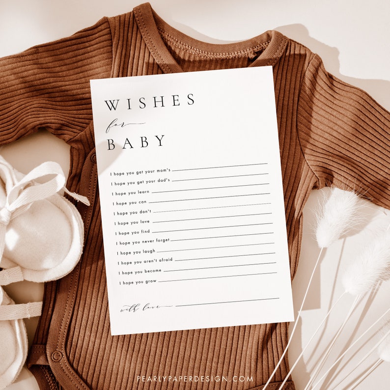 Może przedstawiać: Biała karta z czarnym tekstem, kt&oacute;ry brzmi "Wishes for Baby" i zawiera listę życzeń dla noworodka. Karta leży na brązowym body dla niemowląt i białych butach dla niemowląt.