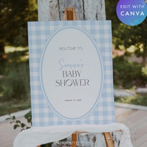 May include: A baby shower sign on an easel. The sign has a light blue gingham pattern background with a white oval in the center. The text reads "WELCOME TO Sienna's BABY SHOWER" and "AUGUST 15, 20XX".
