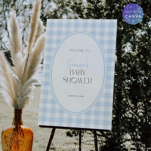 May include: A baby shower sign with a blue and white gingham pattern. The sign reads "Welcome to Sunni's Baby Shower" with the date August 15, 2018. The sign is on a black easel, with a decorative vase and pampas grass in the foreground.