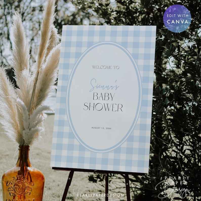 May include: A baby shower sign with a blue and white gingham pattern. The sign reads "Welcome to Sunni's Baby Shower" with the date August 15, 2018. The sign is on a black easel, with a decorative vase and pampas grass in the foreground.