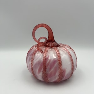 Può includere: Una zucca decorativa in vetro con un corpo bianco e rosa a spirale e un gambo rosso. La zucca ha una superficie strutturata con macchie rosse. Il gambo è arricciato e aggiunge un tocco unico al design.