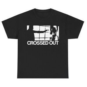 May include: Black t-shirt featuring a white graphic of a silhouetted figure pointing a gun at another figure, set against a window grid. The words "CROSSED OUT" are printed in white below the graphic.