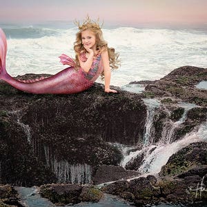 Può includere: Una giovane ragazza vestita da sirena è seduta su uno sperone roccioso vicino all'oceano. Ha una coda rosa e una corona di ramoscelli sulla testa. Le onde dell'oceano si infrangono sullo sfondo.