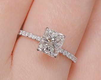 May include: A silver ring with a large, square-shaped diamond set in the center. The diamond is surrounded by smaller diamonds that form a halo around it. The ring is set on a thin band.