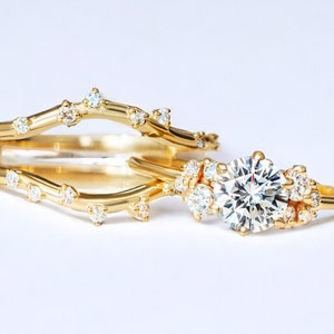 May include: A delicate gold ring set with a large, round, clear diamond surrounded by smaller diamonds. The ring is set on a thin band with a delicate, twig-like design. The ring is paired with a matching band with a similar twig-like design.