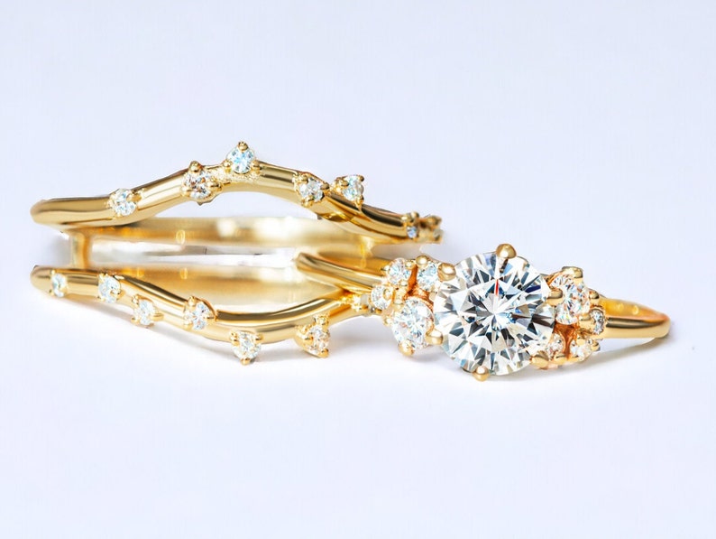 May include: A delicate gold ring set with a large, round, clear diamond surrounded by smaller diamonds. The ring is set on a thin band with a delicate, twig-like design. The ring is paired with a matching band with a similar twig-like design.