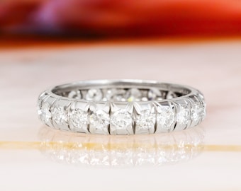 May include: A platinum eternity ring with a continuous row of round-cut diamonds. The diamonds are set closely together, creating a sparkling band. The ring is displayed on a reflective surface, highlighting its brilliance.