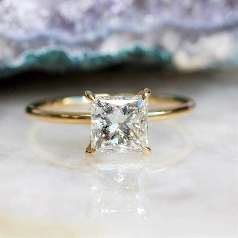Puede incluir: Un anillo de compromiso de banda dorada con un gran diamante cuadrado transparente.