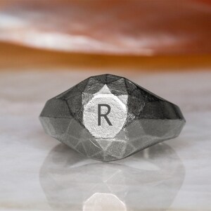 Op de afbeelding: Een zilveren ring met een geometrisch ontwerp en de letter "R" gegraveerd op de bovenkant.