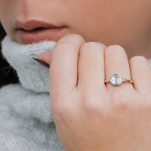 Puede incluir: Un anillo de oro con un diamante de talla esmeralda y dos diamantes baguette m&aacute;s peque&ntilde;os en la banda.