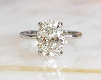 May include: A brilliant oval-cut diamond ring with a silver band. The diamond is the focal point, showcasing its facets and sparkle. The ring is set on a white and beige marble surface, reflecting the light.
