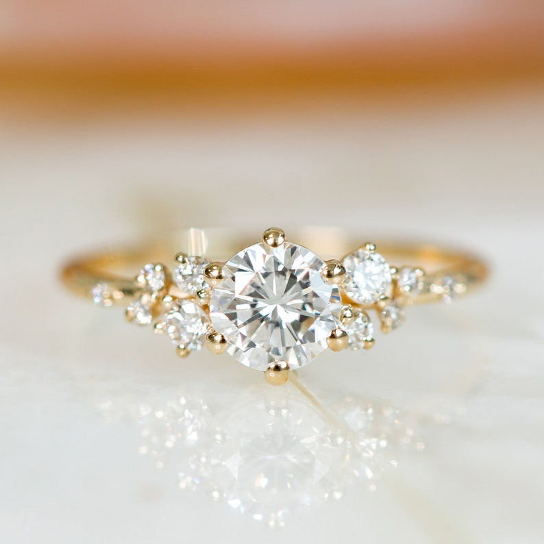 May include: A gold engagement ring with a large round center diamond and smaller diamonds set around it in a cluster design.