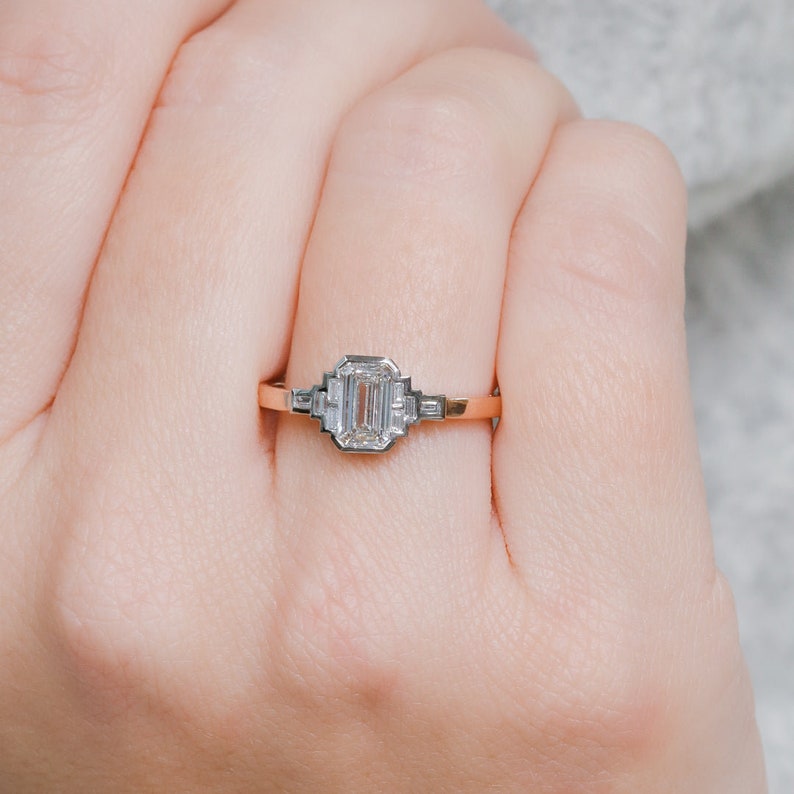 Puede incluir: Un anillo de compromiso de oro rosa con un diamante de talla esmeralda y diamantes m&aacute;s peque&ntilde;os en la banda.